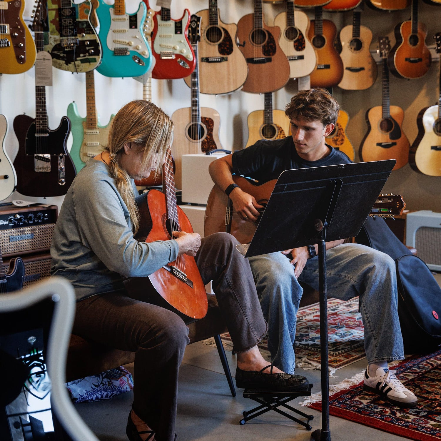 Two people playing guitars during a lesson in a music store with guitars on the wall.