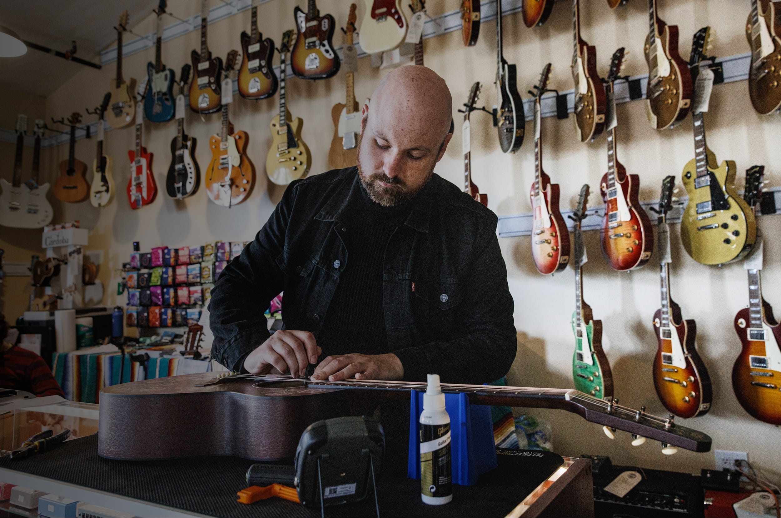 Man working on a guitar in a music store with guitars hanging on the wall.